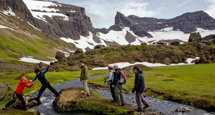 People hiking across a stream in a mountainous snowy landscape.