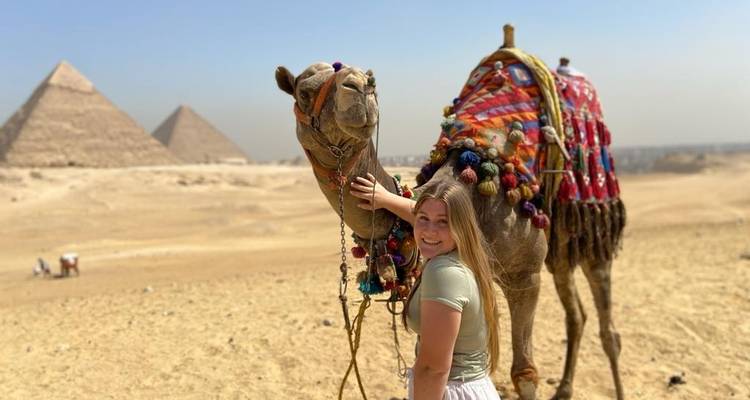Une personne caressant un chameau dans le désert avec des pyramides en arrière-plan.