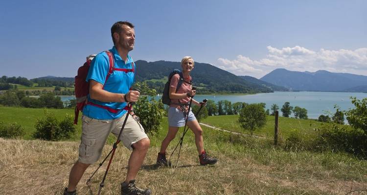 Zwei Personen wandern auf einem Weg in der Nähe eines Sees mit Bergen im Hintergrund.