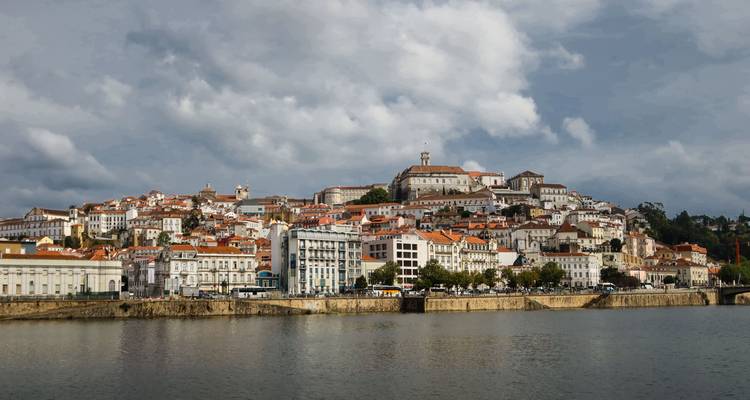 Panorama de la ligne d'horizon riveraine de Coimbra avec des bâtiments aux toits orange sous un ciel dramatique.