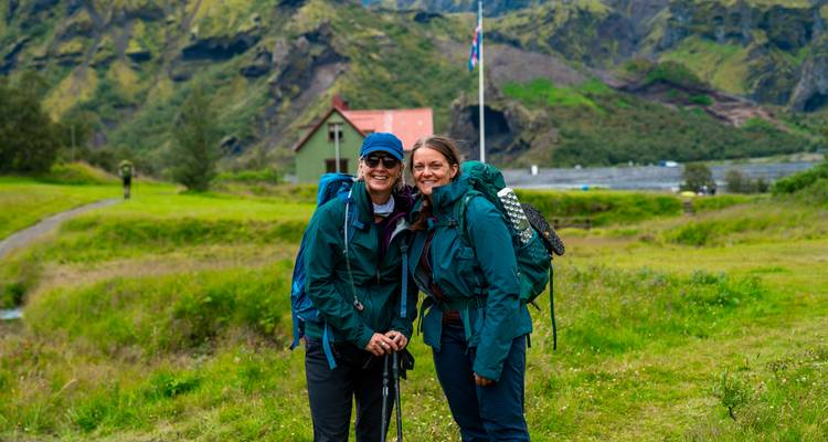 Deux femmes souriantes vêtues d'équipements de trekking se tiennent debout dans une prairie herbeuse avec un refuge de montagne en arrière-plan
