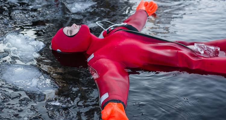 Person in einem roten Überlebensanzug schwimmt ruhig zwischen Eisbrocken in dunklem arktischen Wasser.