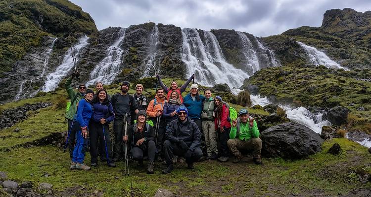 Large hiking group celebrating in front of a multi-tier waterfall cascading down mossy cliffs