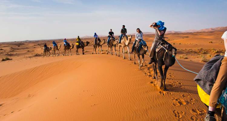 Caravana de camellos cruzando dunas del desierto con personas sentadas sobre camellos.