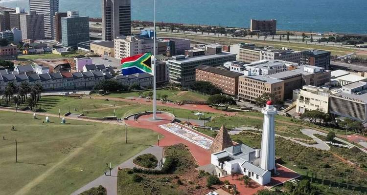 Vista aérea de la ciudad con una enorme bandera sudafricana en un mástil alto junto a un parque urbano costero y un faro.