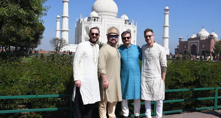 Four men wearing traditional Indian kurtas pose in front of the white marble Taj Mahal on a sunny day.