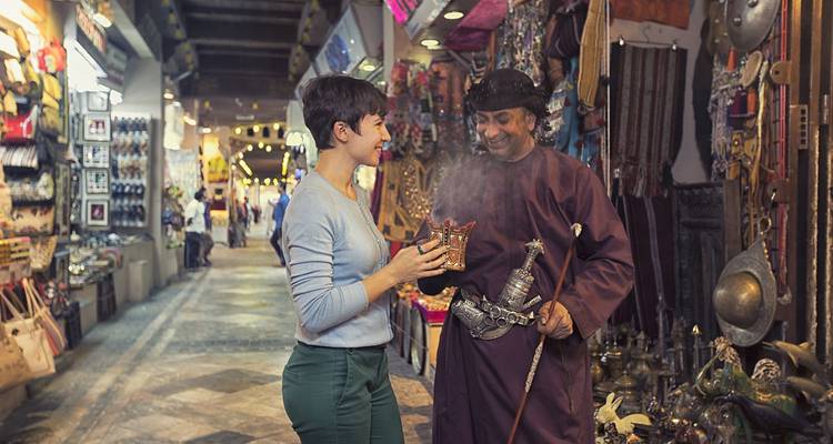 Mercado interior con una persona interactuando con un comerciante, estantes llenos de mercancía.