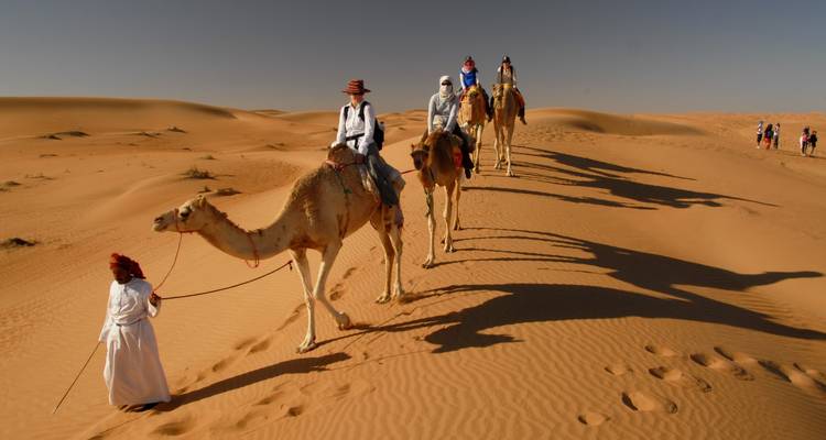 Una fila de camellos con jinetes cruzando dunas del desierto; guía liderando al frente.
