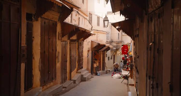 Smalle medina steeg met houten deuren en luifels, lokale verkoper die goederen rangschikt in warm middaglicht