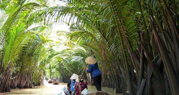 Des gens qui pagayent à travers un passage d'eau étroit entouré de palmiers dans un cadre tropical.