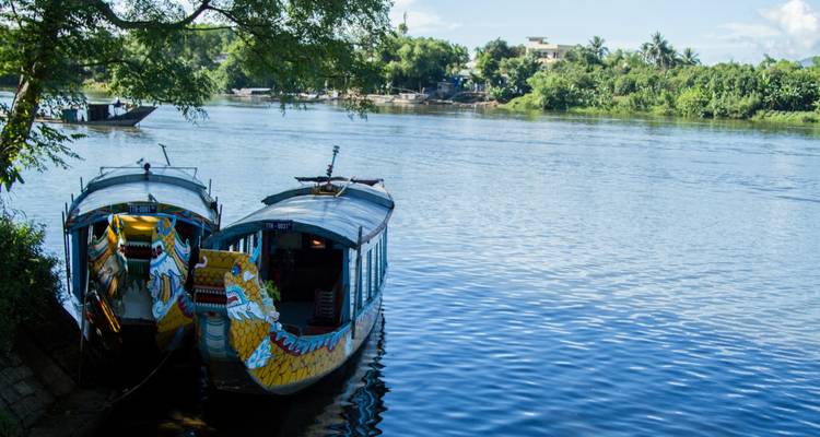 Zwei Drachenboote am Ufer eines Flusses mit Bäumen und Gebäuden im Hintergrund.