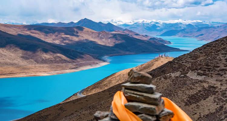 Steinhaufen mit orangefarbenem Gebetsschal behangen überblickt glänzend blauen See und umgebende braune Berge unter fleckigen Wolken.