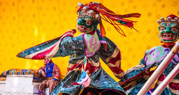 Ein lebhaft maskierter tibetischer Tänzer wirbelt in mehrfarbigen Roben vor einem leuchtend gelben Hintergrund während eines traditionellen Cham-Festivals.