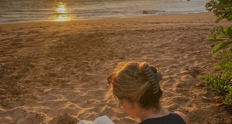 Person reading a book by the beach during sunset.