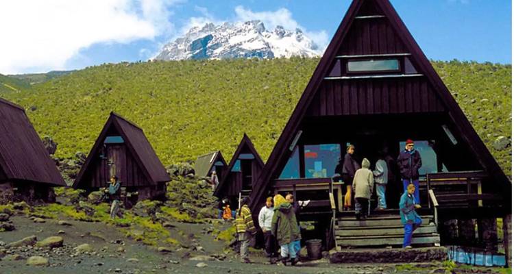 Des huttes en bois de forme triangulaire parsèment une pente volcanique avec le sommet enneigé du Kilimandjaro visible au-delà.