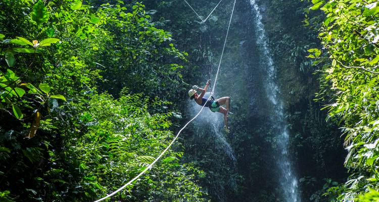 Un canyonista se columpia en una cuerda junto a una alta cascada de la selva, con el spray brillando bajo el sol.