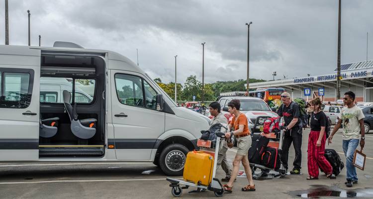 Personas cargando equipaje en una furgoneta blanca estacionada afuera de una terminal de aeropuerto.