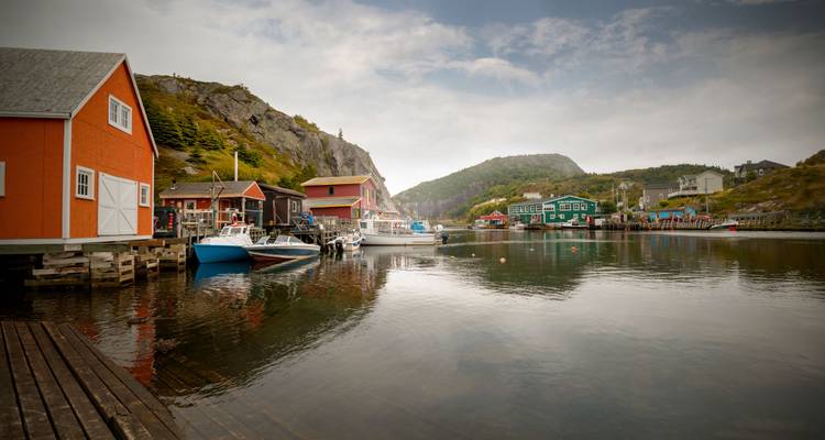 Des maisons en bois colorées et des bateaux de pêche bordent un port tranquille niché entre des collines boisées.