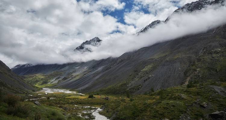 Dramatisch alpien dal met rivier, mistige wolken die tegen steile rotsachtige hellingen hangen en verspreide begroeiing.