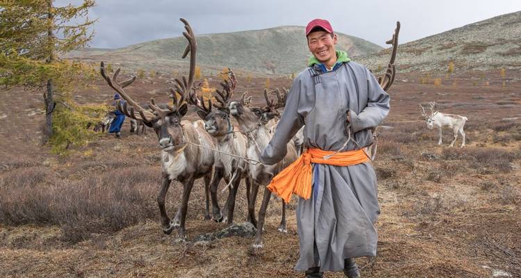 Een glimlachende nomadische herder in traditionele gewaad leidt een kleine groep rendieren over de kale toendra.