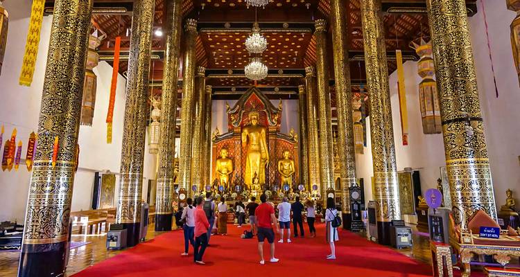 Interior de templo dorado ornamentado con visitantes y una gran estatua de Buda.