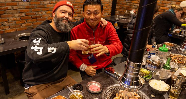Two men toast glasses over sizzling Korean barbecue in a cozy brick-walled restaurant.