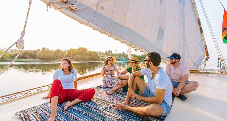 Travelers relaxing on a felucca sailboat deck as it glides along a calm river at sunset