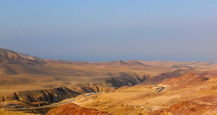 Vast expanse of rolling ochre and red desert hills with a winding road disappearing into haze