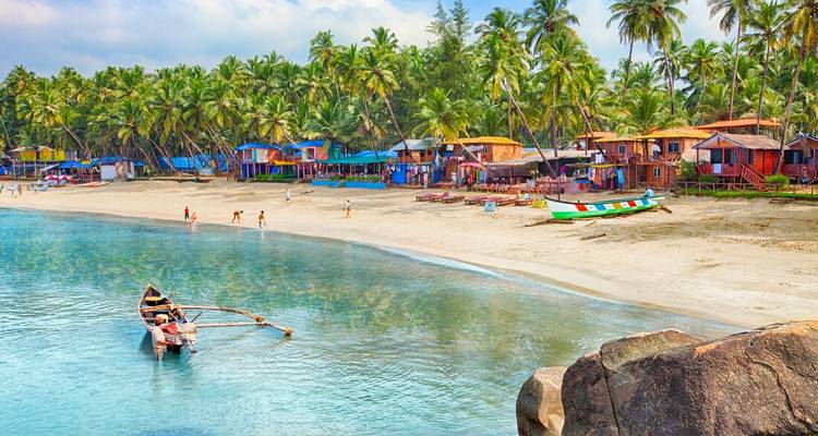 Village coloré et plage de sable bordée de palmiers avec bateau de pêche sur une eau bleue cristalline à Goa