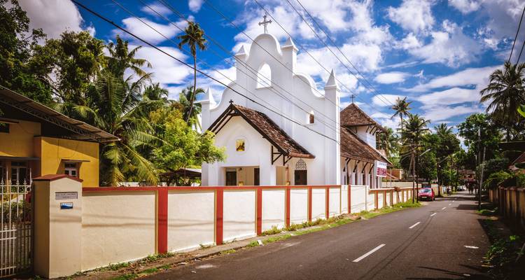 Pequeña iglesia blanca al borde de la carretera con techo de tejas rojas bajo un cielo parcialmente nublado en Kerala.