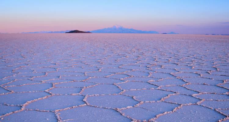 Polígonos de sal agrietada al amanecer con montaña distante bajo un cielo pastel en Uyuni.