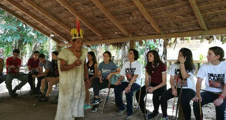 Grupo de turistas sentados bajo un techo de paja escuchando a una persona vestida con atuendo tradicional.