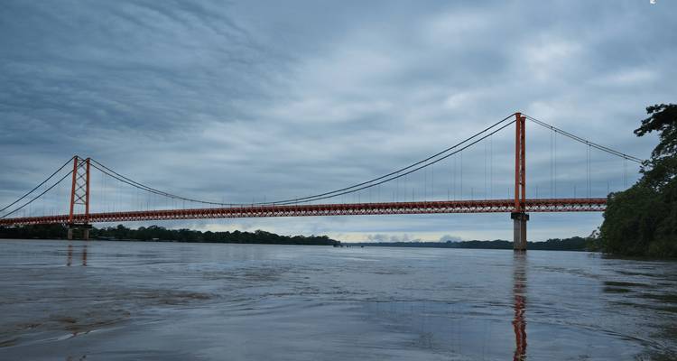 Puente colgante rojo y largo que cruza un río Amazonas ancho bajo un cielo nublado
