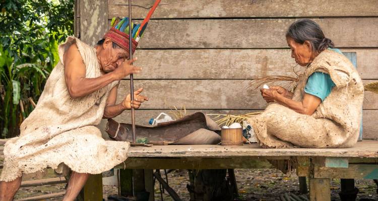 Dos personas indígenas en traje tradicional elaborando objetos mientras están sentadas en una plataforma de madera fuera de una casa en la selva