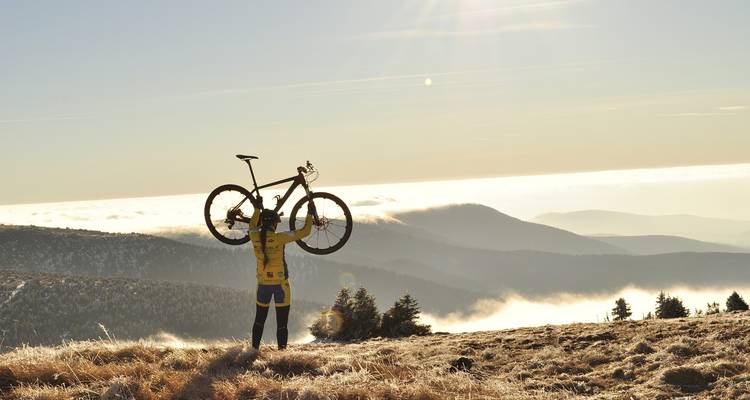 Persona sosteniendo una bicicleta sobre su cabeza con un fondo escénico de montañas.
