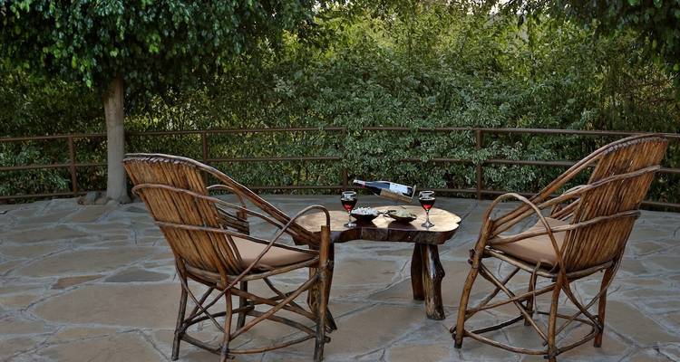 Dos sillas de madera y una mesa con bebidas en un patio de piedra, rodeado de naturaleza.