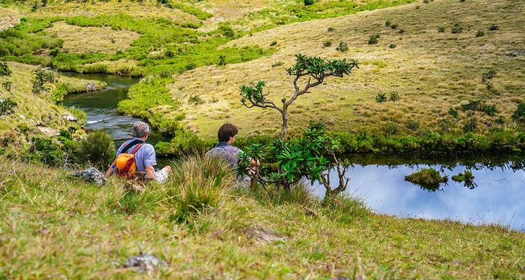 Two people sitting by a stream in a hilly landscape.