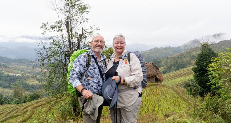 Couple âgé souriant avec des sacs à dos posant devant des rizières en terrasses luxuriantes et des collines brumeuses.