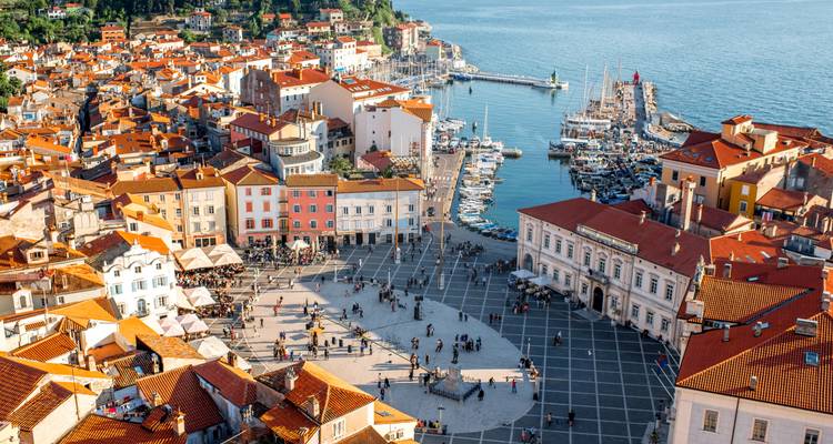 Vue panoramique d'une ville côtière méditerranéenne avec des toits rouges entourant une marina et une vaste place centrale.