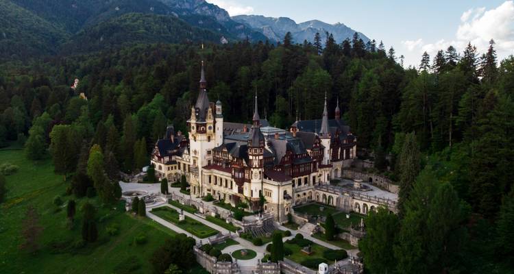 Vue aérienne du château de Peles niché dans une épaisse forêt de conifères avec les pics des Carpates en arrière-plan.