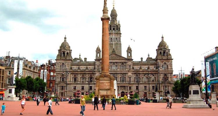 George Square à Glasgow avec la majestueus City Chambers derrière de imposants monuments et des personnes qui traversent la plaza.