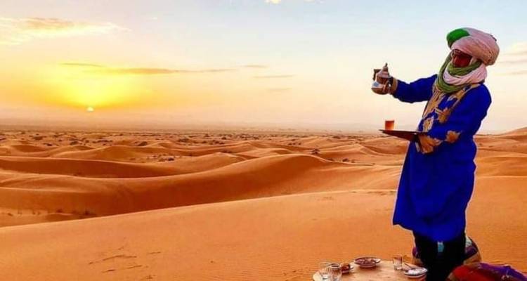 Man in traditionele kledij die thee zet in de Saharawoestijn tijdens zonsondergang.