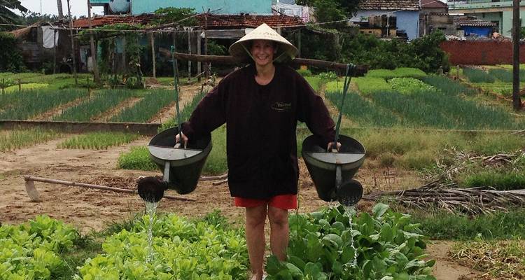 Un visitante con un sombrero cónico riega filas de verduras vibrantes en un huerto rural.