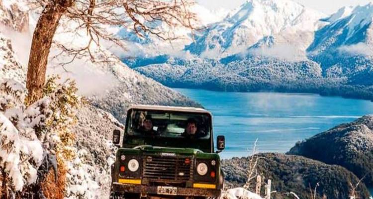 Las montañas cubiertas de nieve se alzan sobre un lago azul mientras un 4x4 verde navega por un camino de invierno accidentado
