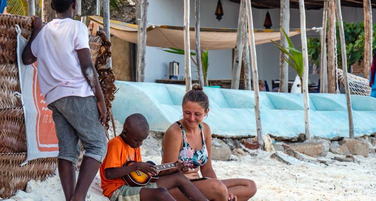 Boy playing a musical instrument with a woman sitting beside him on the beach.