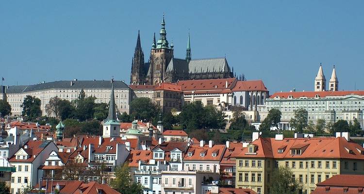 Panoramic view of Prague Castle and St. Vitus Cathedral rising above red-roofed houses under a clear blue sky.