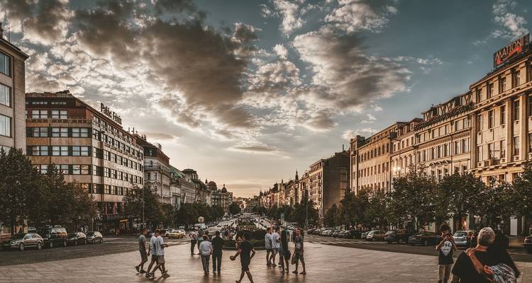Wide-angle sunset view down Wenceslas Square with pedestrians crossing a broad boulevard framed by historic façades and dramatic clouds.