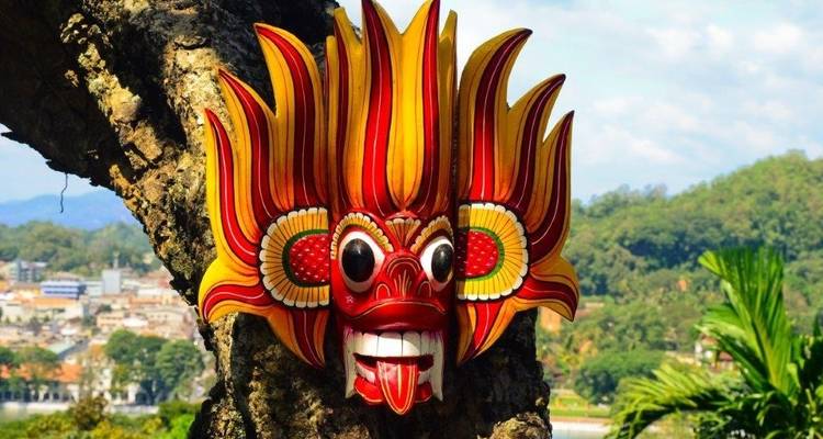 Masque traditionnel sri-lankais suspendu à un arbre avec un paysage urbain en arrière-plan.