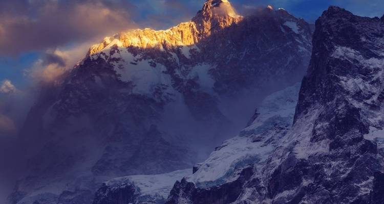 Sonnenbeschienene Gipfel hoher schneebedeckter Berge unter einem dramatischen Himmel.