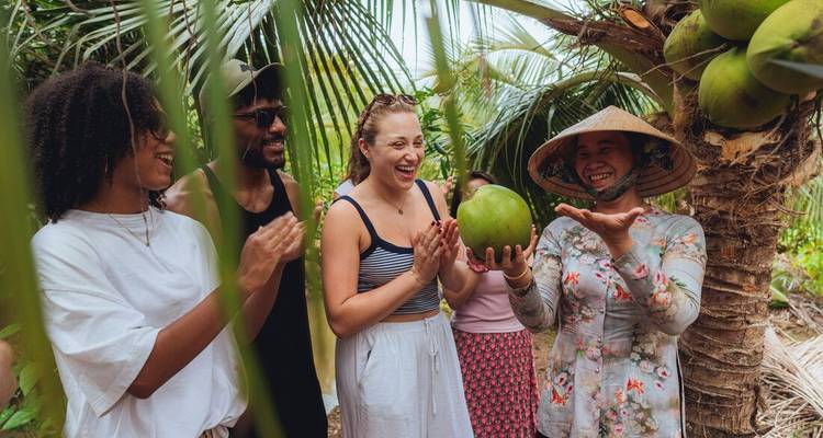 Los visitantes aplauden mientras una mujer local presenta un coco fresco bajo las frondas de palmera.